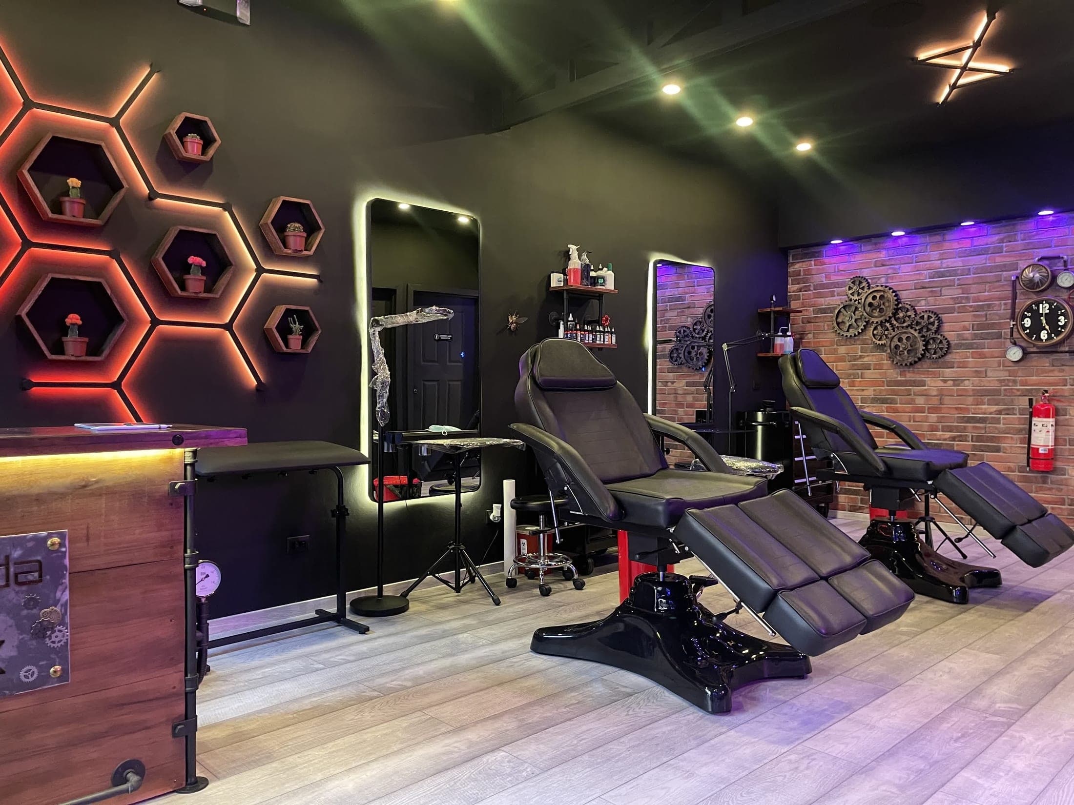 Interior del estudio La Vida Ink: paredes oscuras, estantes hexagonales iluminados, pared de ladrillo con engranajes y dos sillas de tatuaje